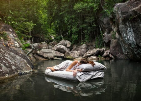 Floating mattress in a serene river surrounded by rocks and lush green trees for outdoor sleeping comfort.
