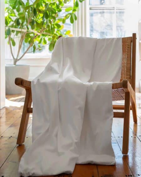 Luxury white bedsheet draped over wooden chair near bright window with green plant in background - mattress and sleep essentials for a healthy mattress-buying experience.